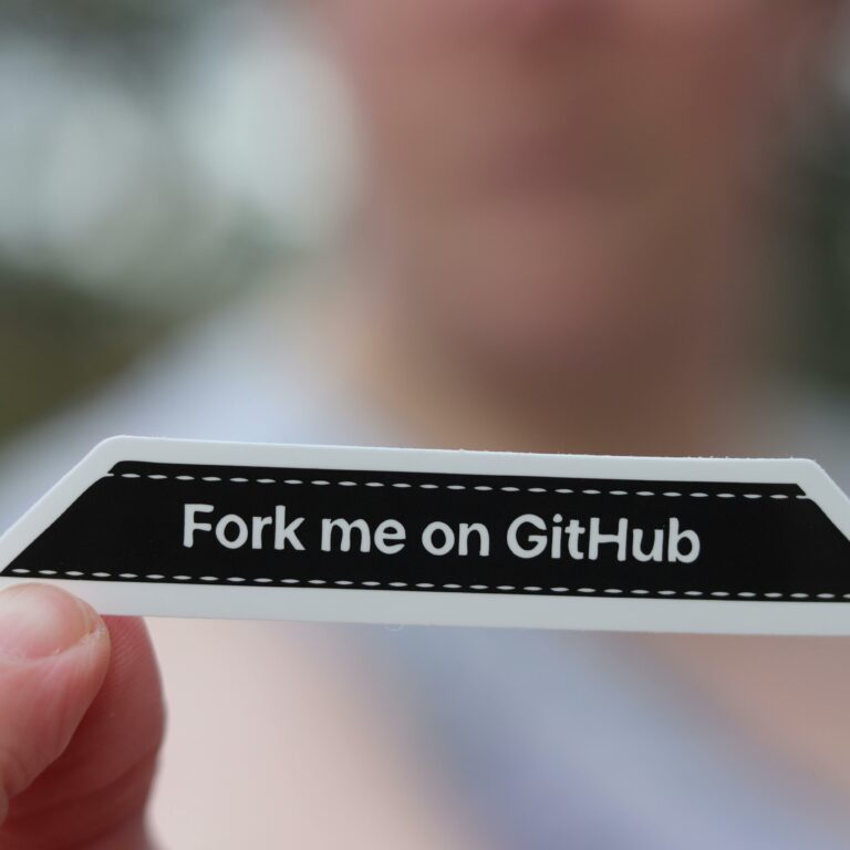Close-up of a hand holding a 'Fork me on GitHub' sticker, blurred background.
